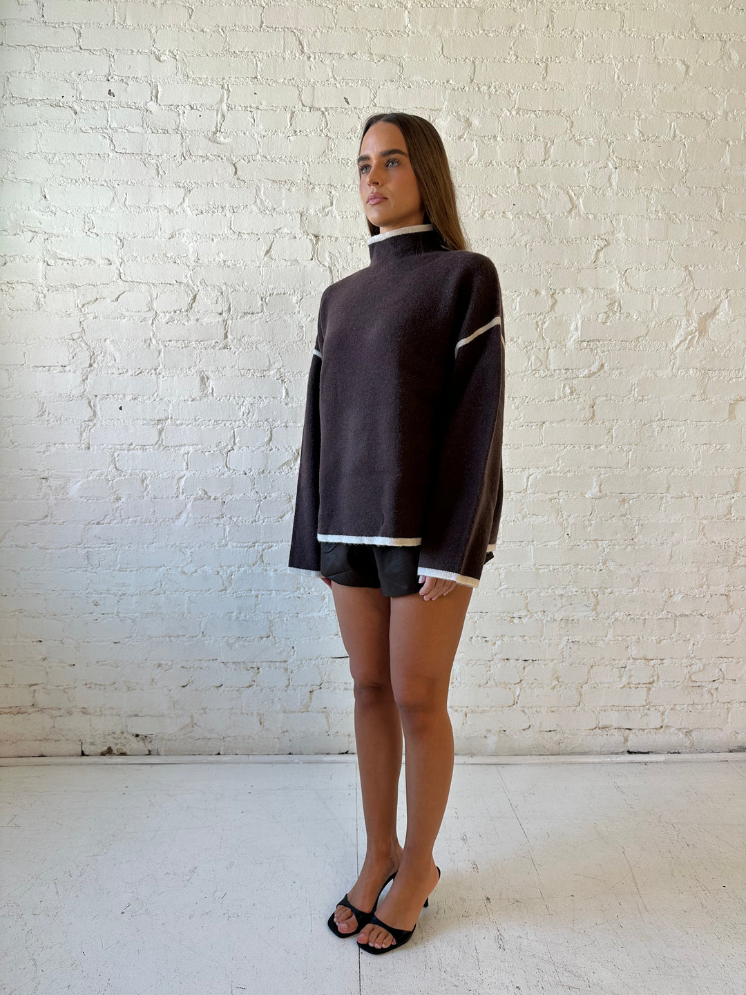 Woman wearing boxy brown oversized sweater with contrast trim and brown leather short. 