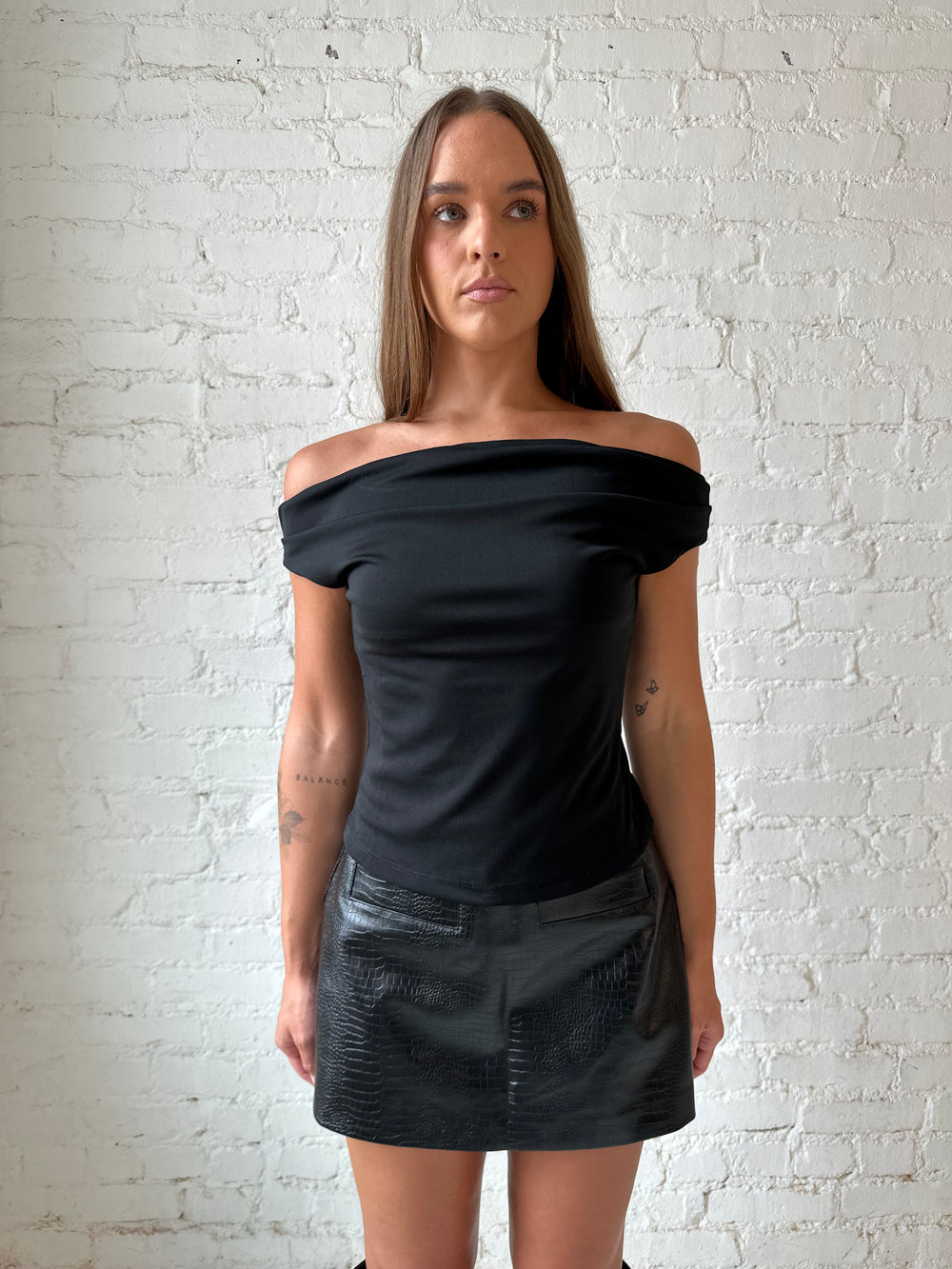 Woman wearing black croc faux leather mini skort with black off the shoulder top, and black knee boots.