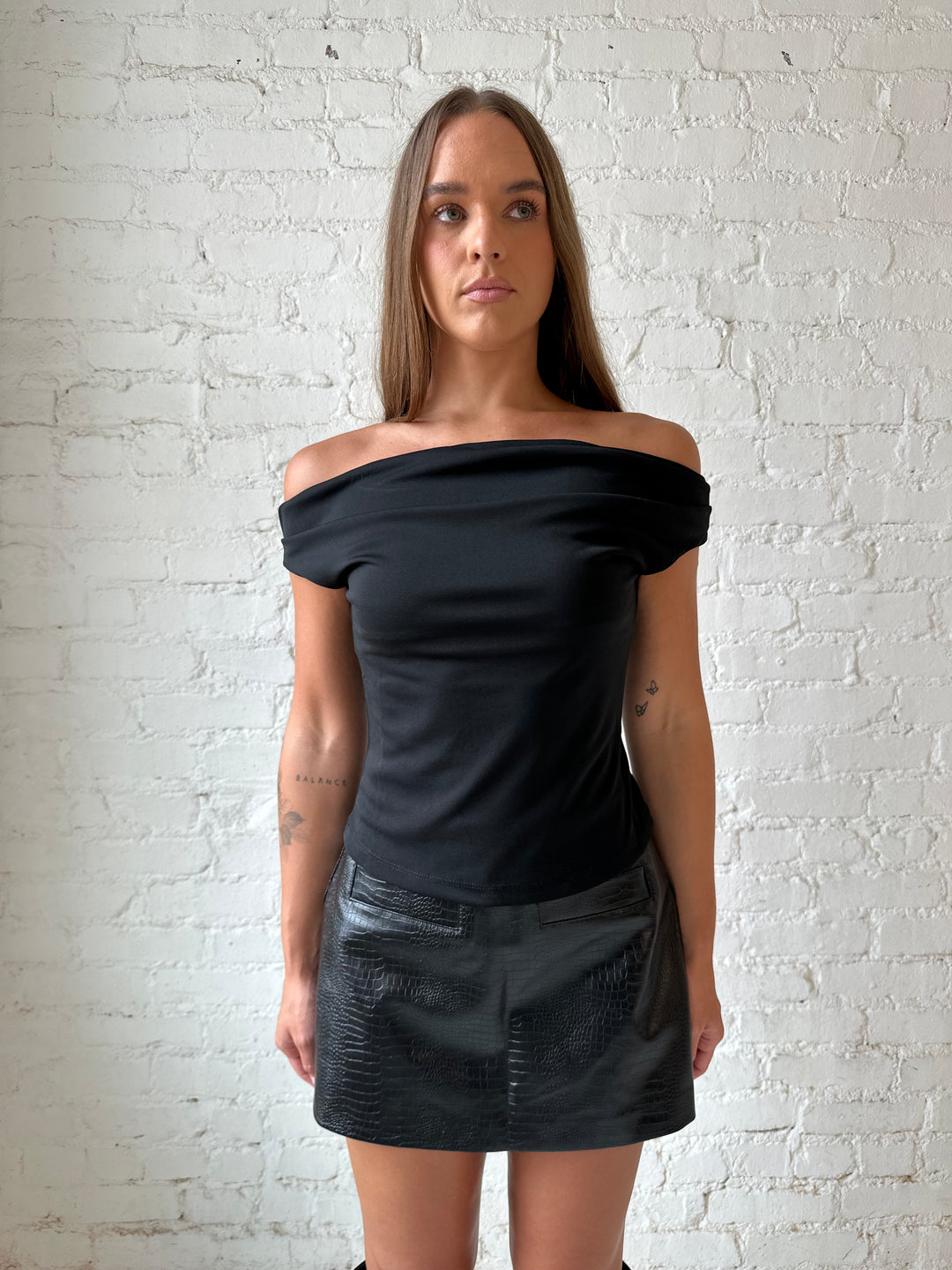 Woman wearing black croc faux leather mini skort with black off the shoulder top, and black knee boots.