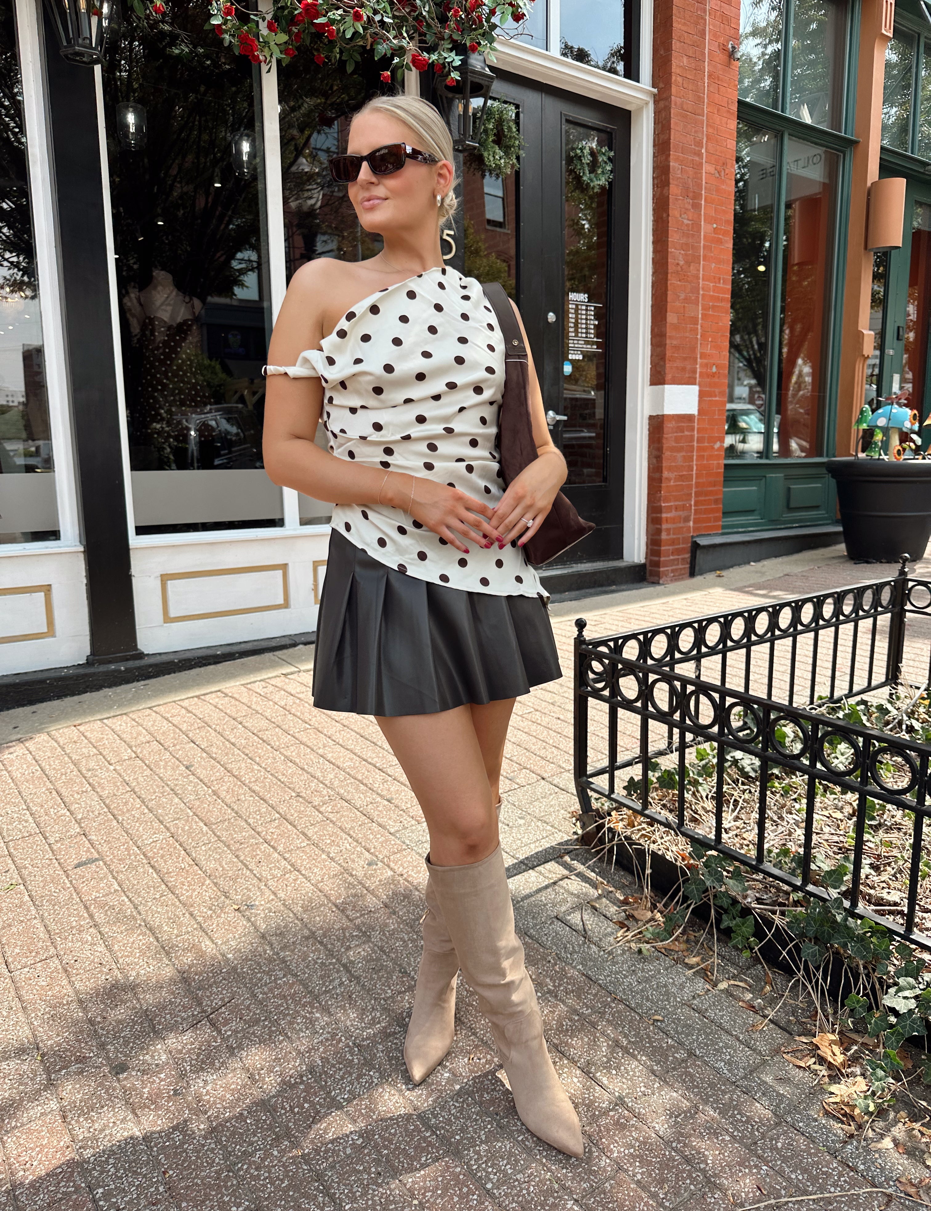 Woman wearing one shoulder satin polka dot top with brown leather pleated mini skirt. 
