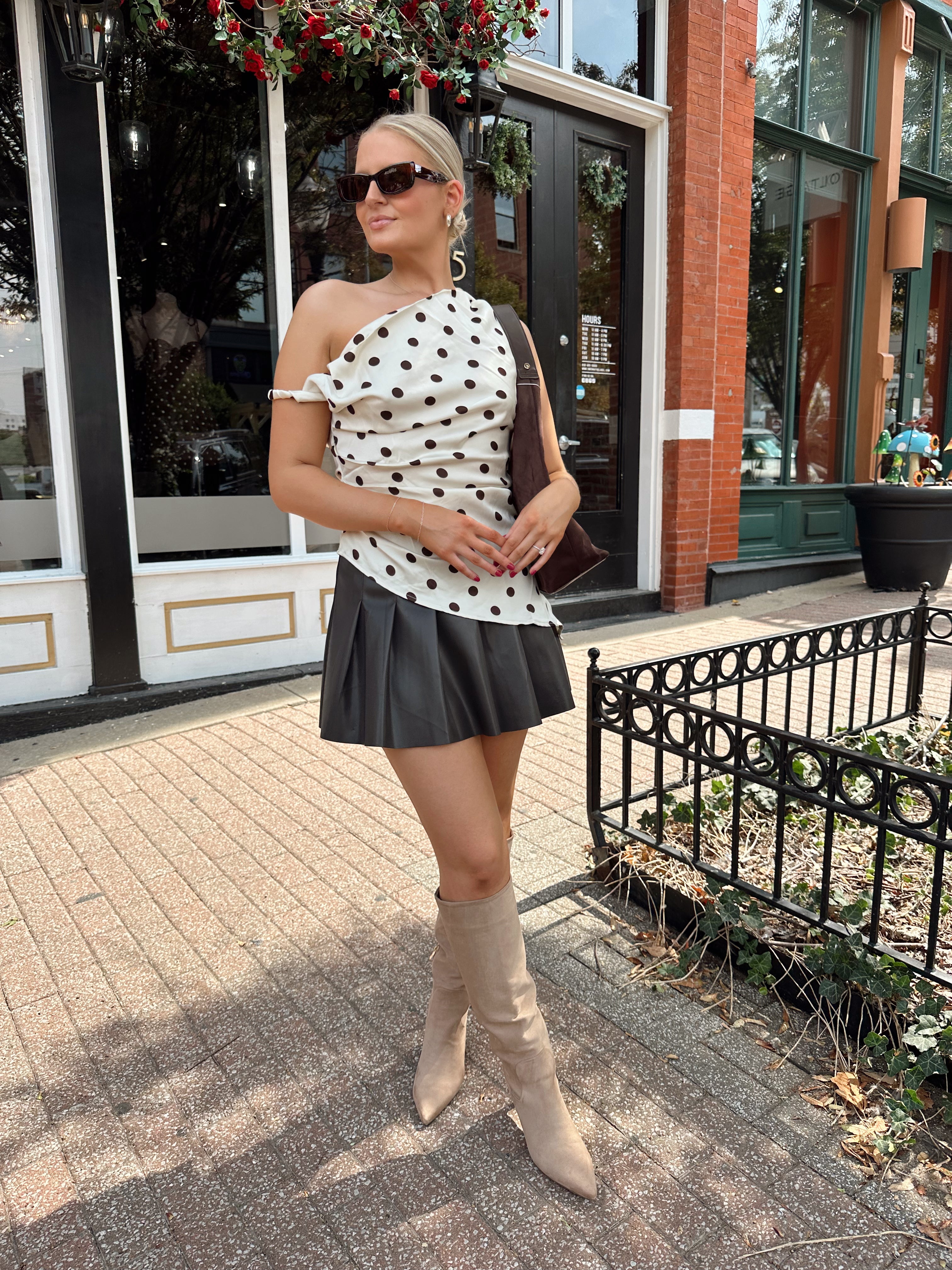 Woman wearing one shoulder satin polka dot top with brown leather pleated mini skirt. 