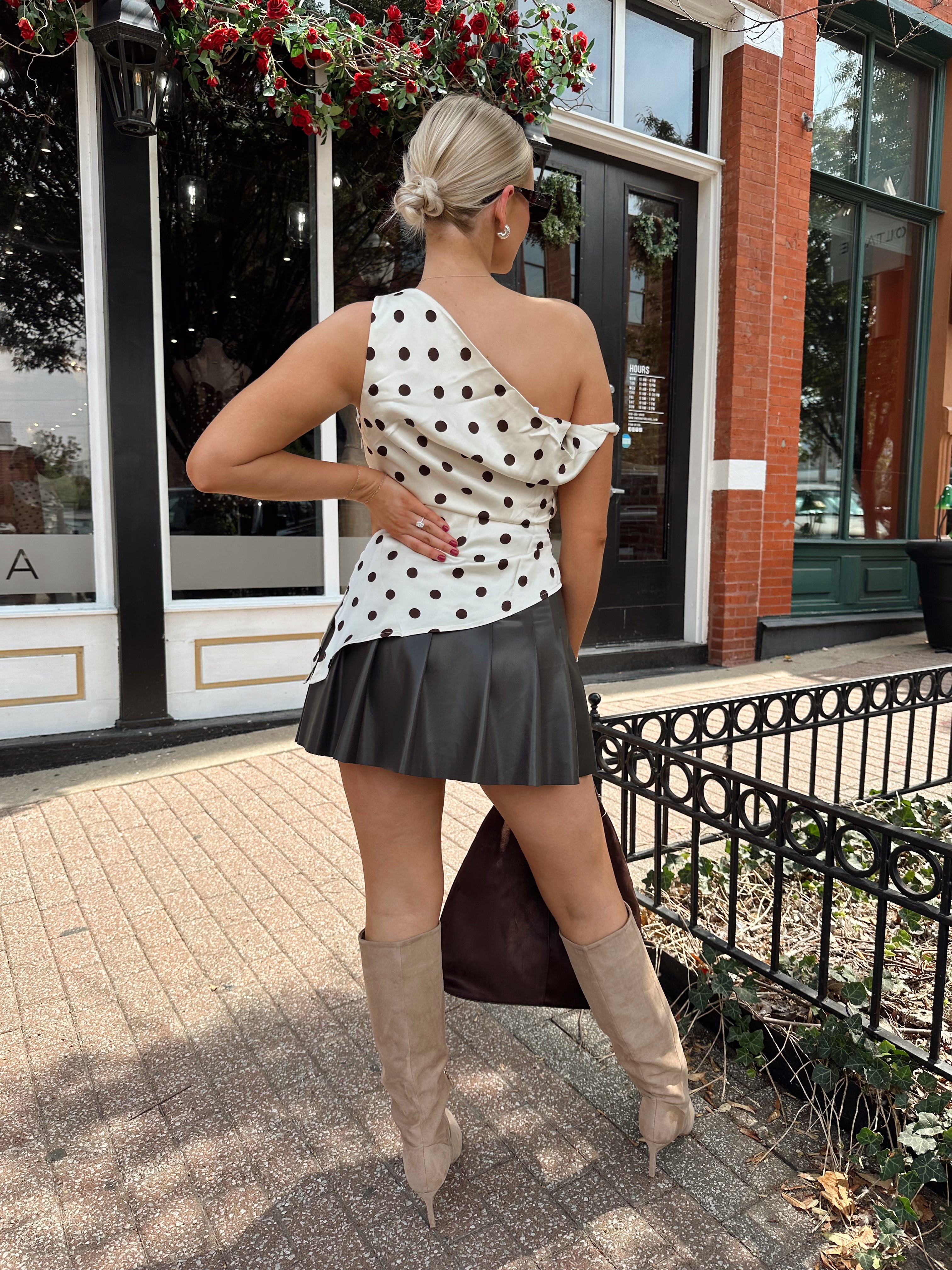 Woman wearing one shoulder satin polka dot top with brown leather pleated mini skirt. 
