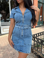 Woman wearing light wash denim mini dress with belt detail and collar neckline.