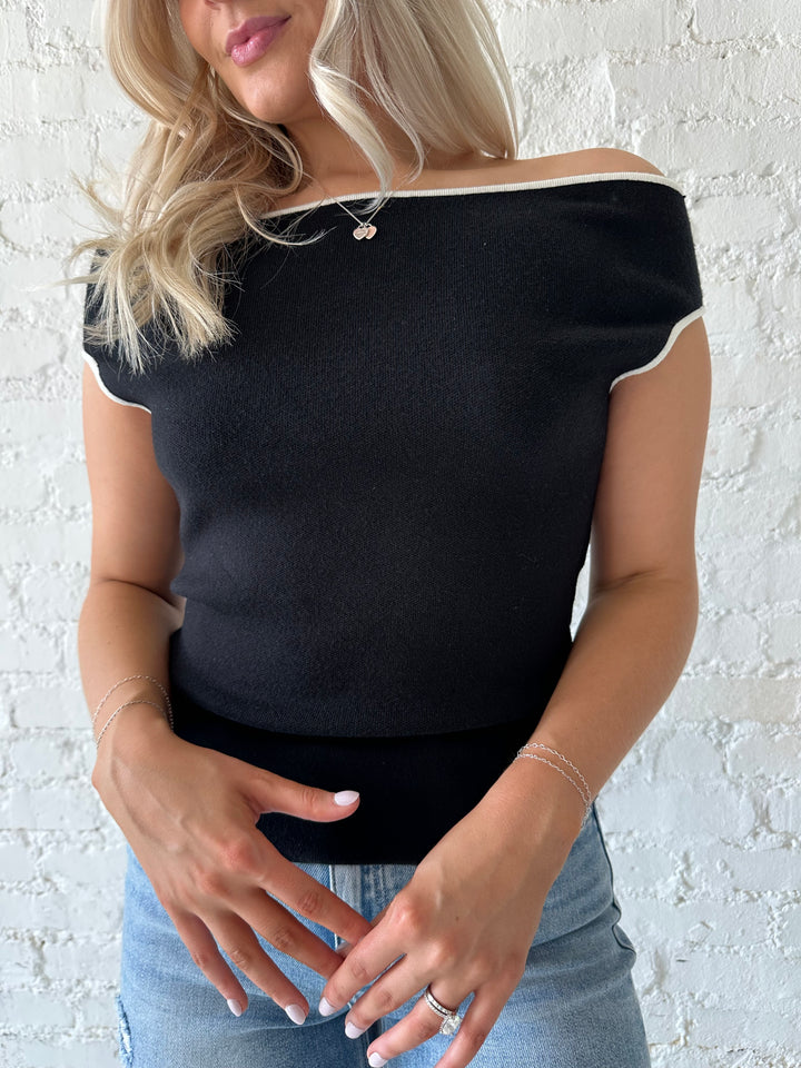Woman wearing black knit off the shoulder top with contrast white trim, and distressed jeans. 