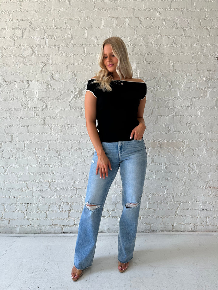 Woman wearing black knit off the shoulder top with white contrast trim and distressed jeans.