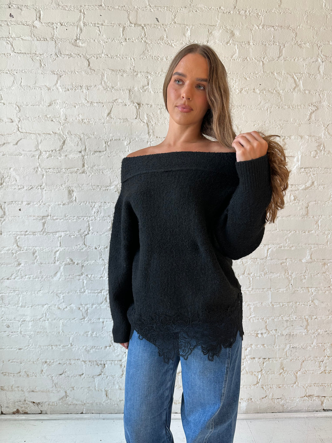 Woman wearing black knit, off the shoulder sweater with lace trim hem, and dark wash jean.