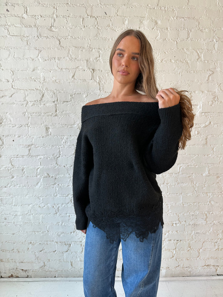 Woman wearing black knit, off the shoulder sweater with lace trim hem, and dark wash jean.