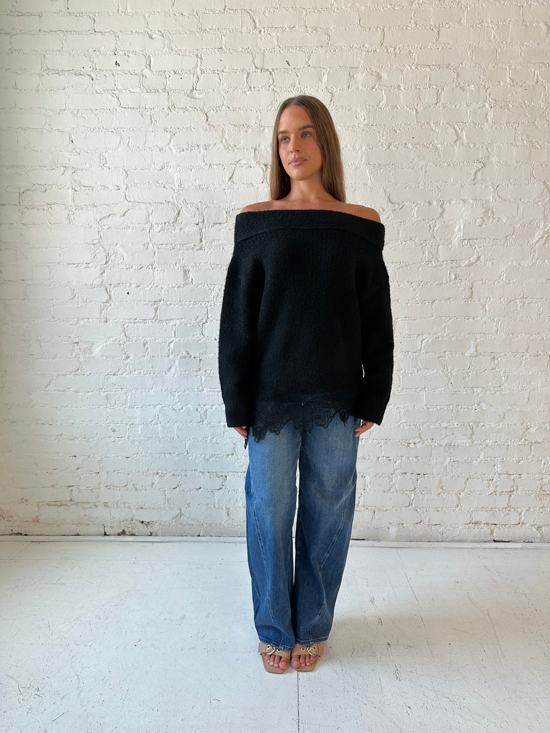 Woman wearing black knit, off the shoulder sweater with lace trim hem, and dark wash jean.