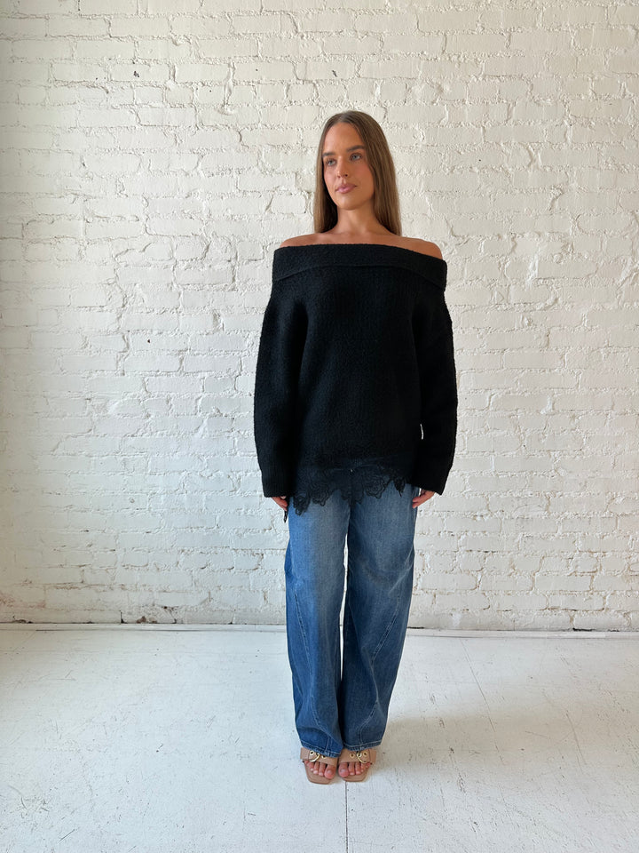 Woman wearing black knit, off the shoulder sweater with lace trim hem, and dark wash jean.