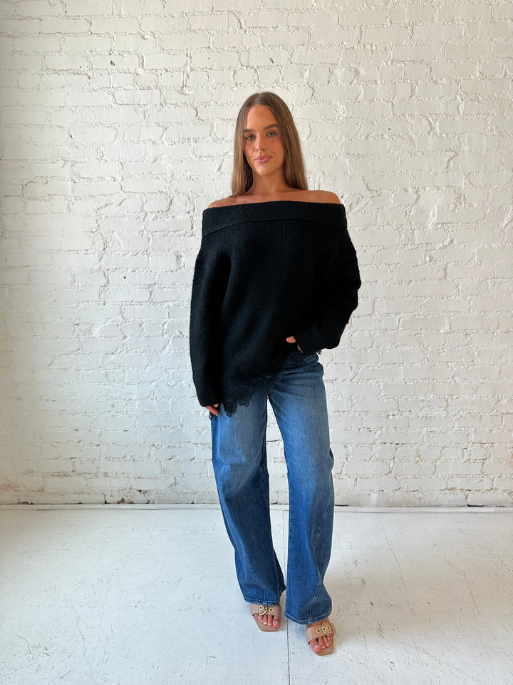 Woman wearing black knit, off the shoulder sweater with lace trim hem, and dark wash jean.
