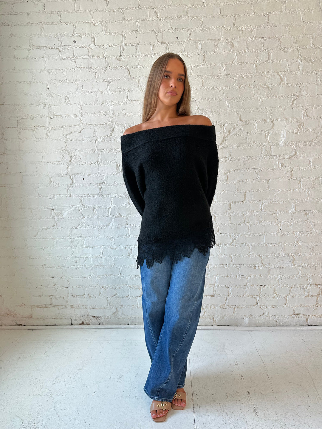 Woman wearing black knit, off the shoulder sweater with lace trim hem, and dark wash jean.