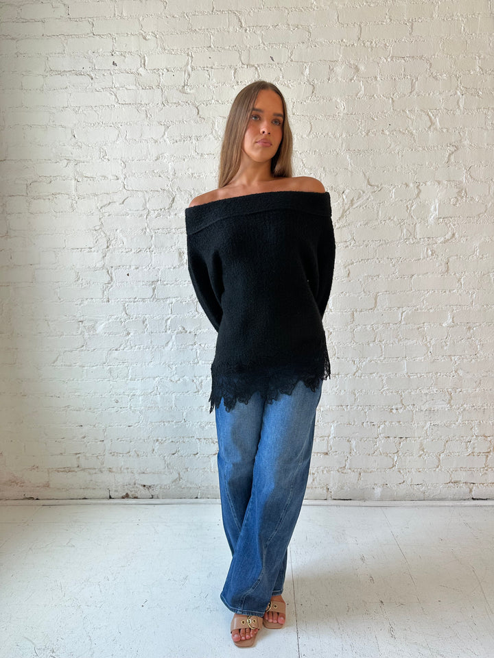 Woman wearing black knit, off the shoulder sweater with lace trim hem, and dark wash jean.