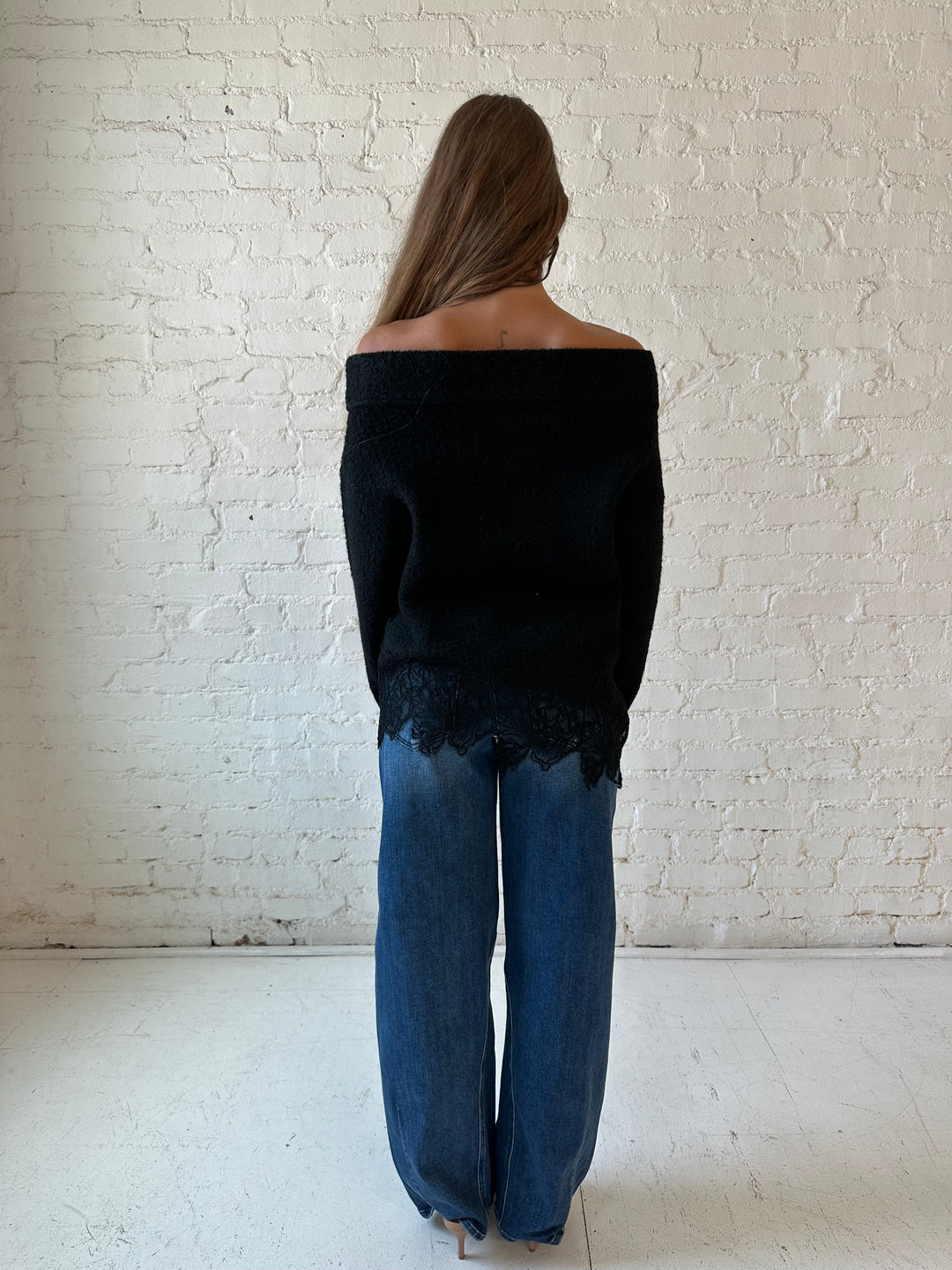 Woman wearing black knit, off the shoulder sweater with lace trim hem, and dark wash jean.