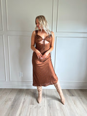 Woman wearing brown satin polka dot midi dress with cutout midsection, lace trim and tie detail.
