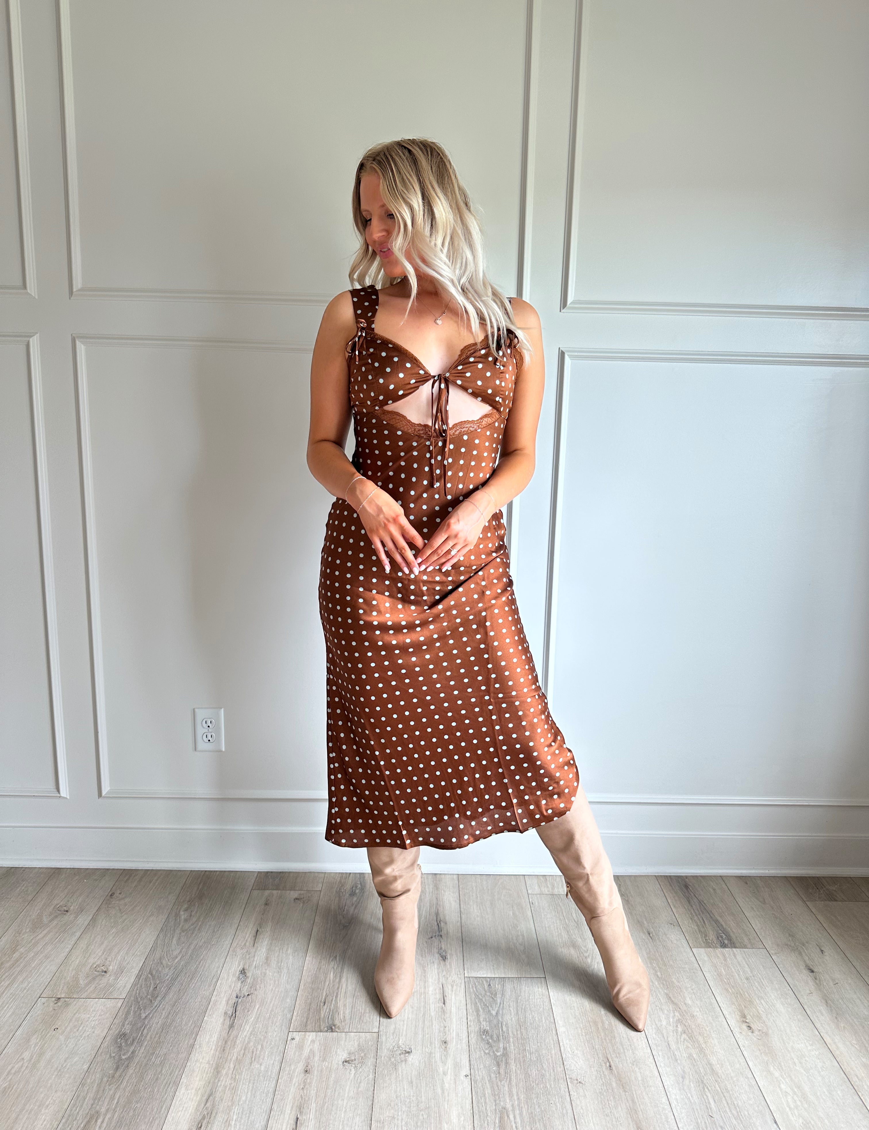 Woman wearing brown satin polka dot midi dress with cutout midsection, lace trim and tie detail.