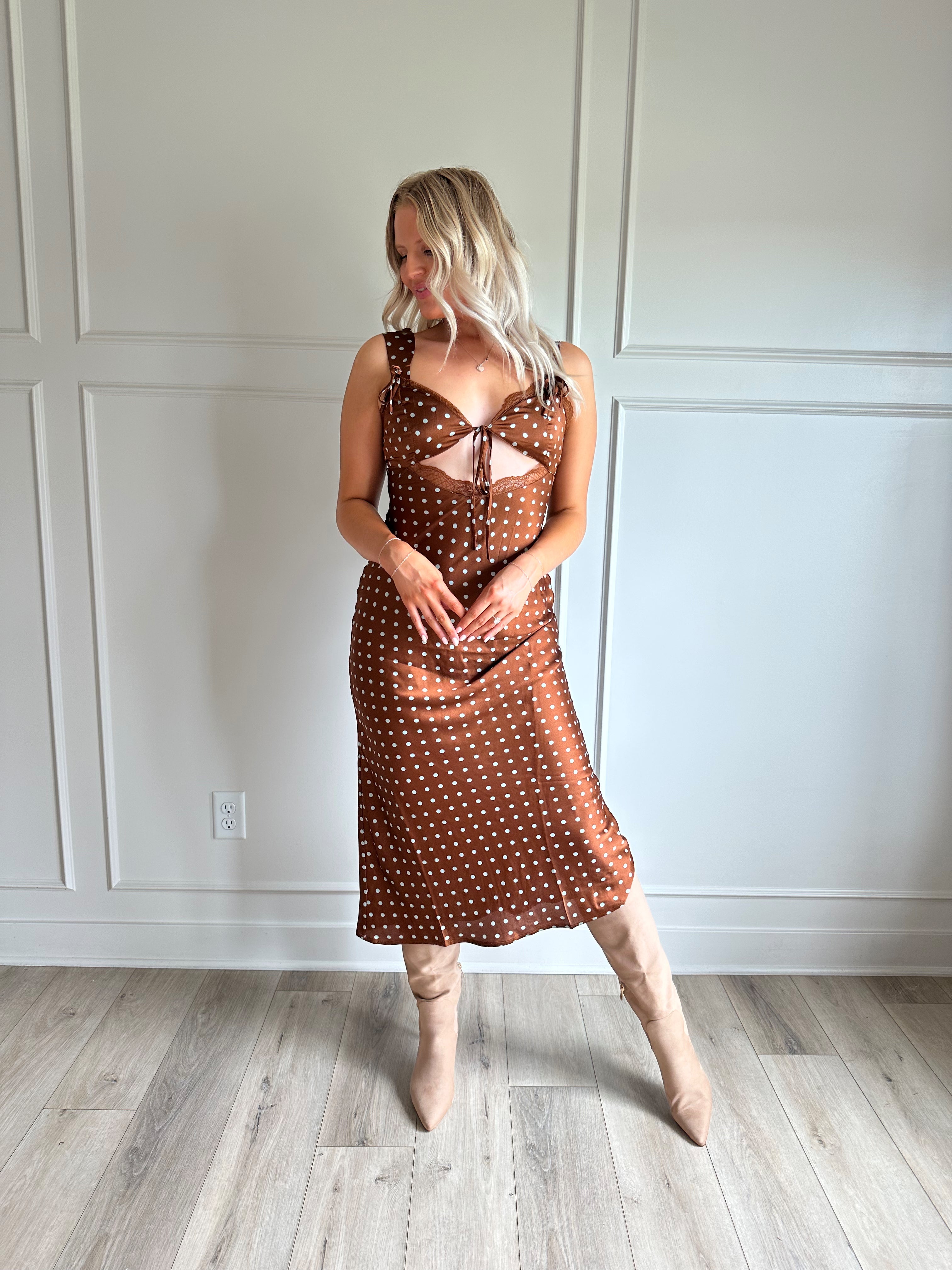 Woman wearing brown satin polka dot midi dress with cutout midsection, lace trim and tie detail.