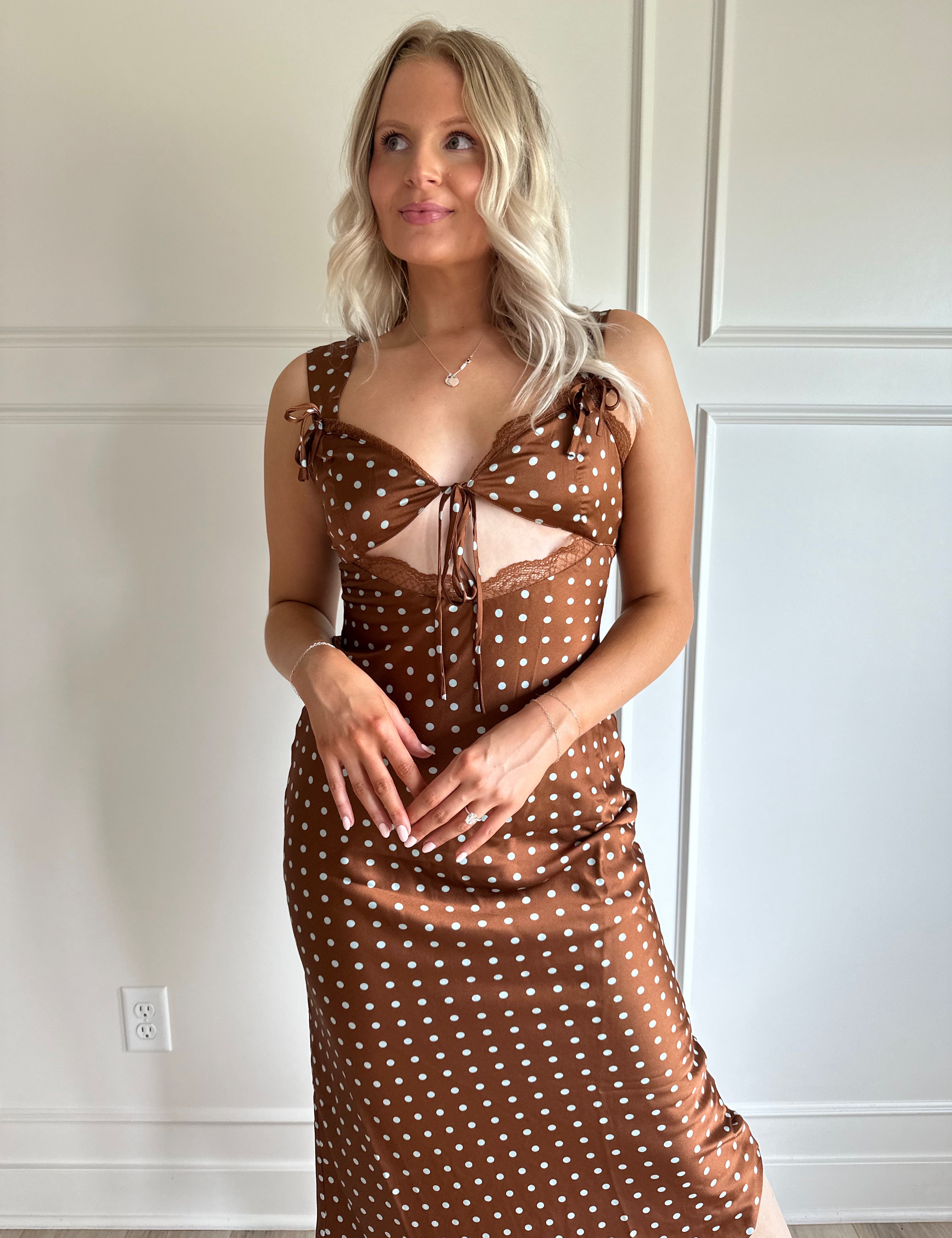 Woman wearing brown satin polka dot midi dress with cutout midsection, lace trim and tie detail.