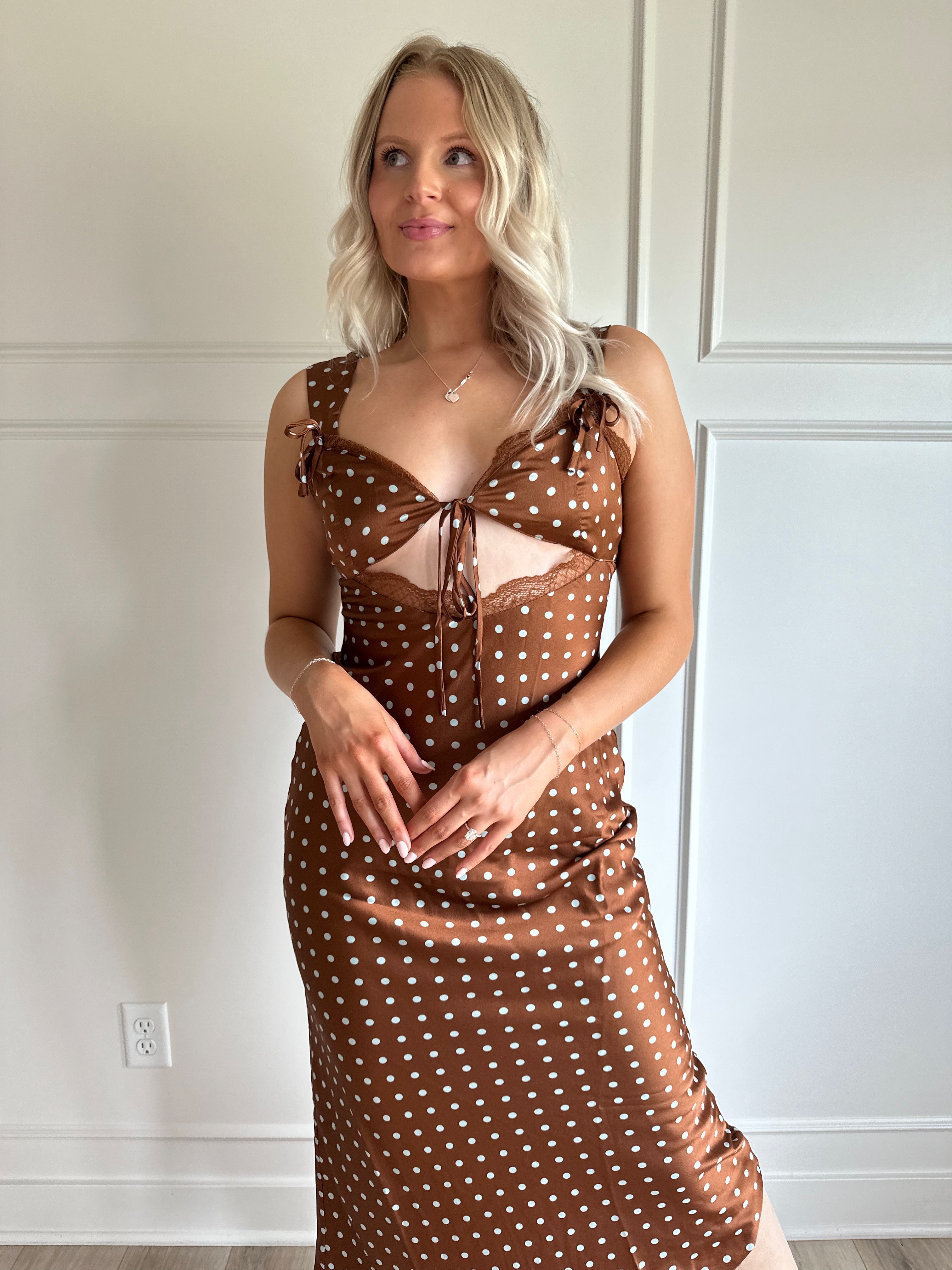 Woman wearing brown satin polka dot midi dress with cutout midsection, lace trim and tie detail.