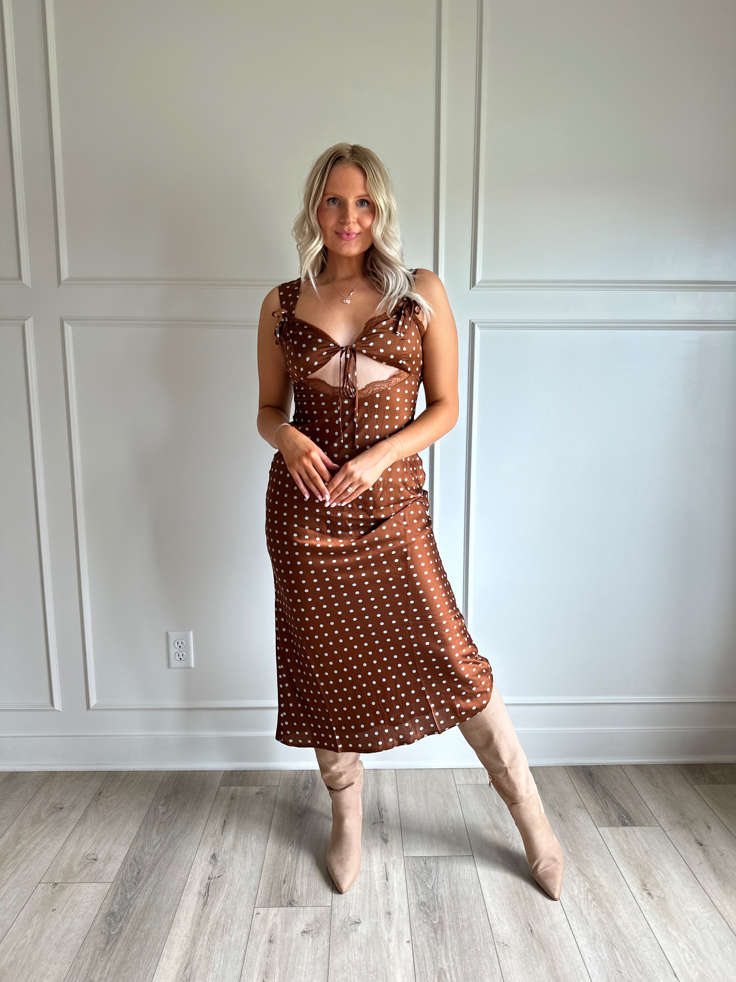 Woman wearing brown satin polka dot midi dress with cutout midsection, lace trim and tie detail.