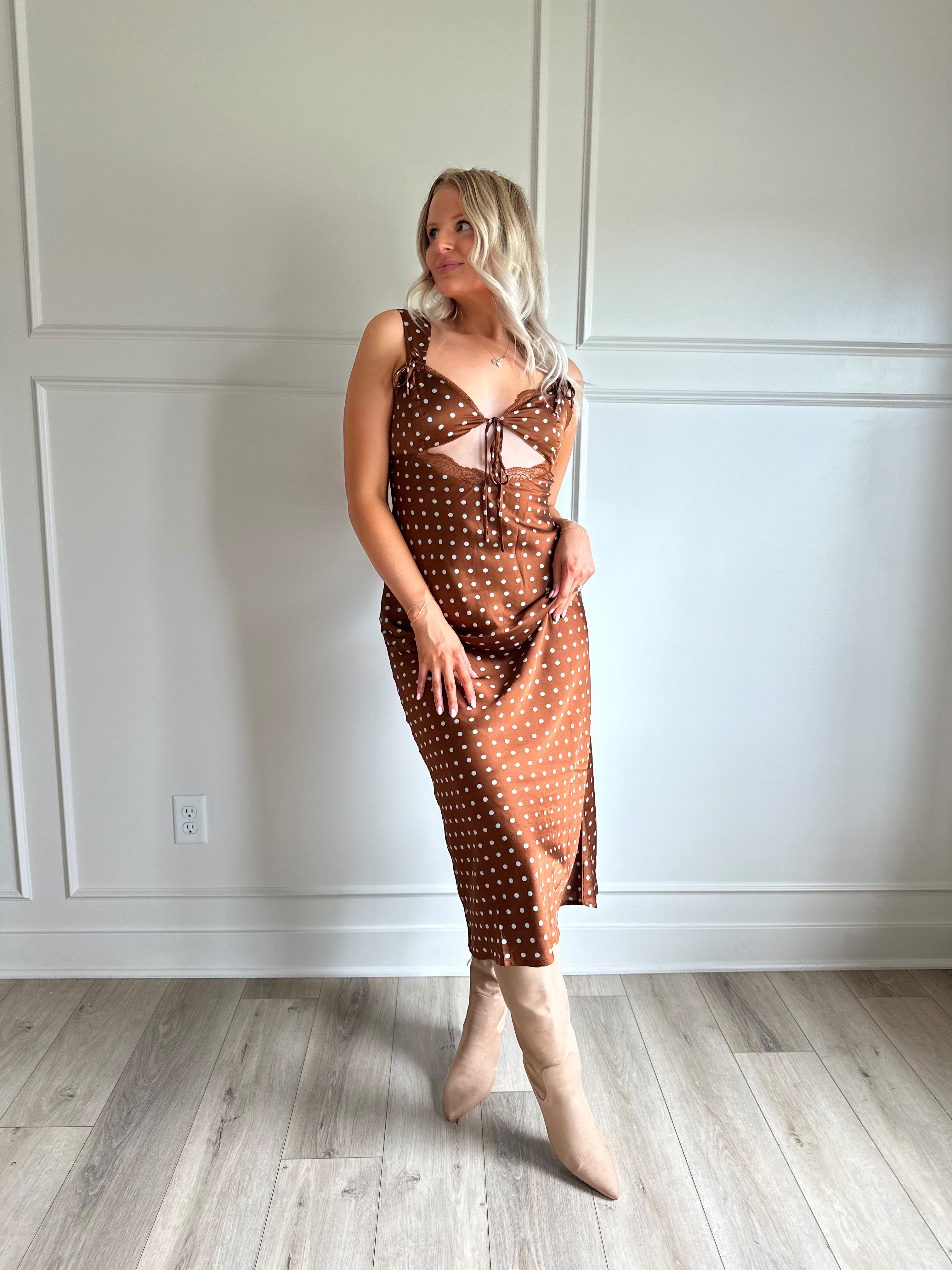 Woman wearing brown satin polka dot midi dress with cutout midsection, lace trim and tie detail.