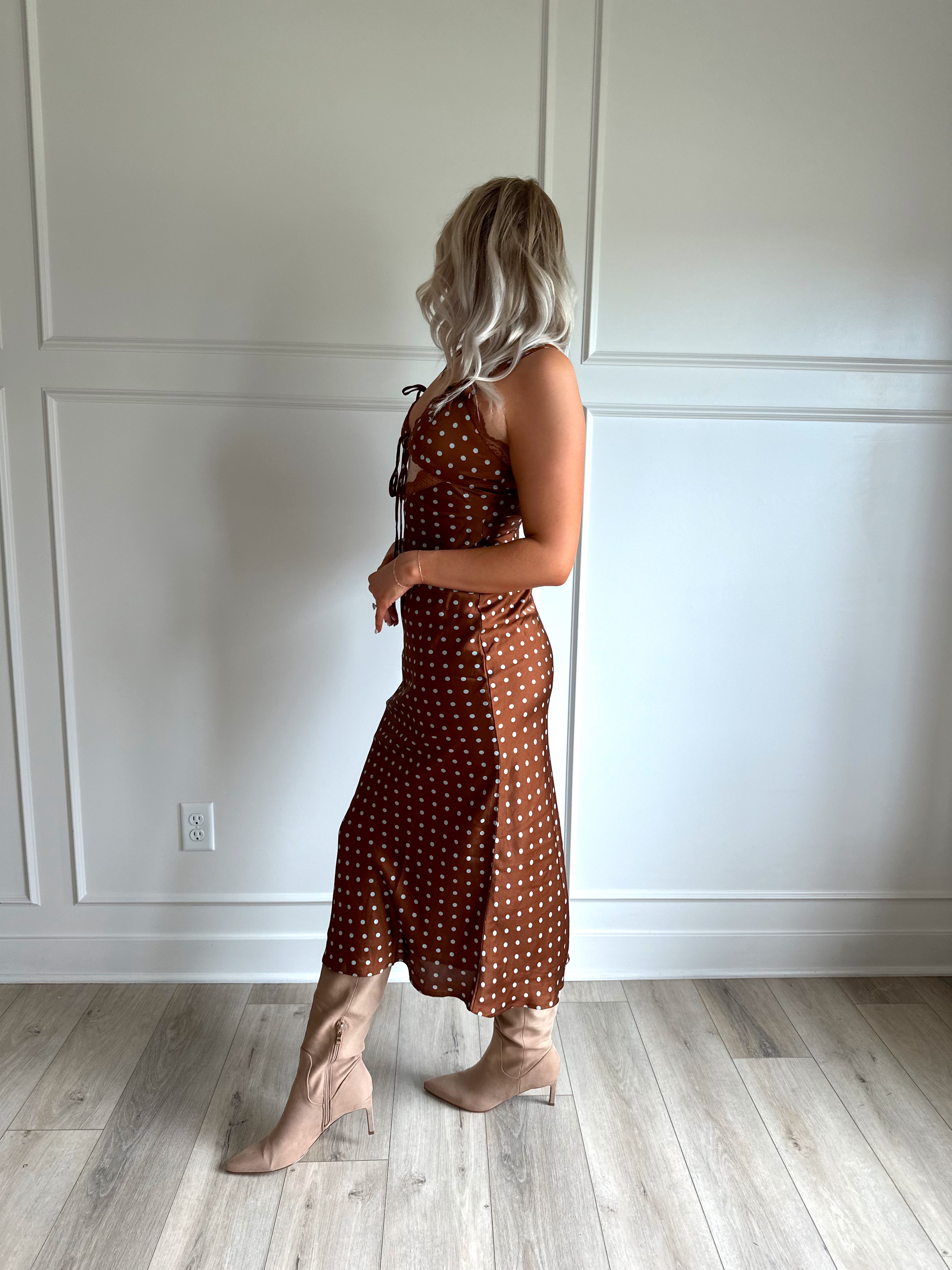 Woman wearing brown satin polka dot midi dress with cutout midsection, lace trim and tie detail.