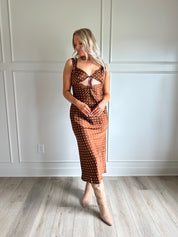 Woman wearing brown satin polka dot midi dress with cutout midsection, lace trim and tie detail.