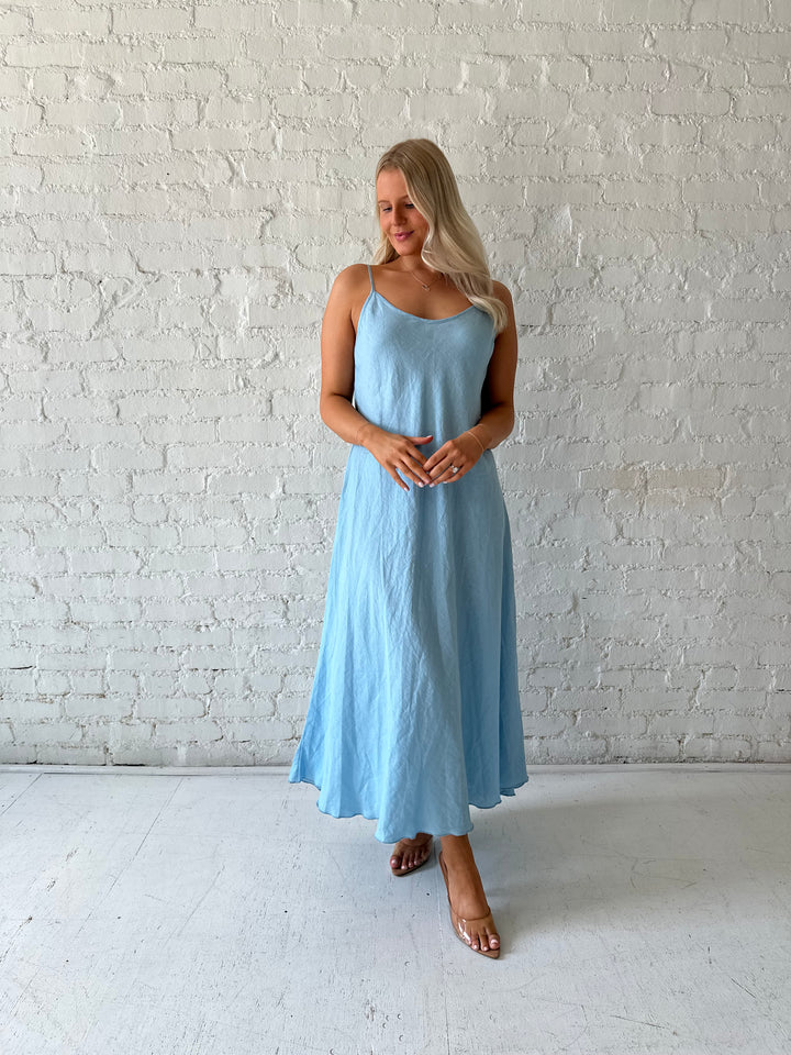 Woman wearing light blue linen slip maxi dress with adjustable spaghetti straps. 