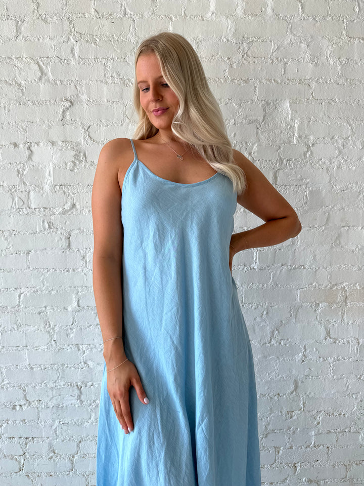 Woman wearing light blue linen slip maxi dress with adjustable spaghetti straps. 