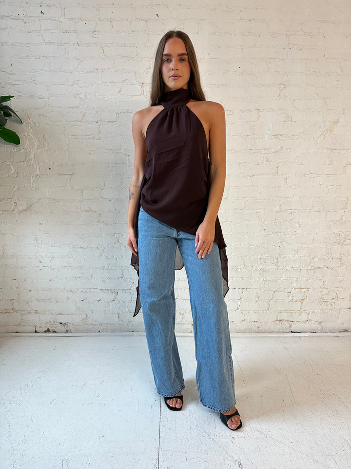 Woman wearing brown chiffon backless top with self tie halter, and frilly detail, and medium wash jeans. 