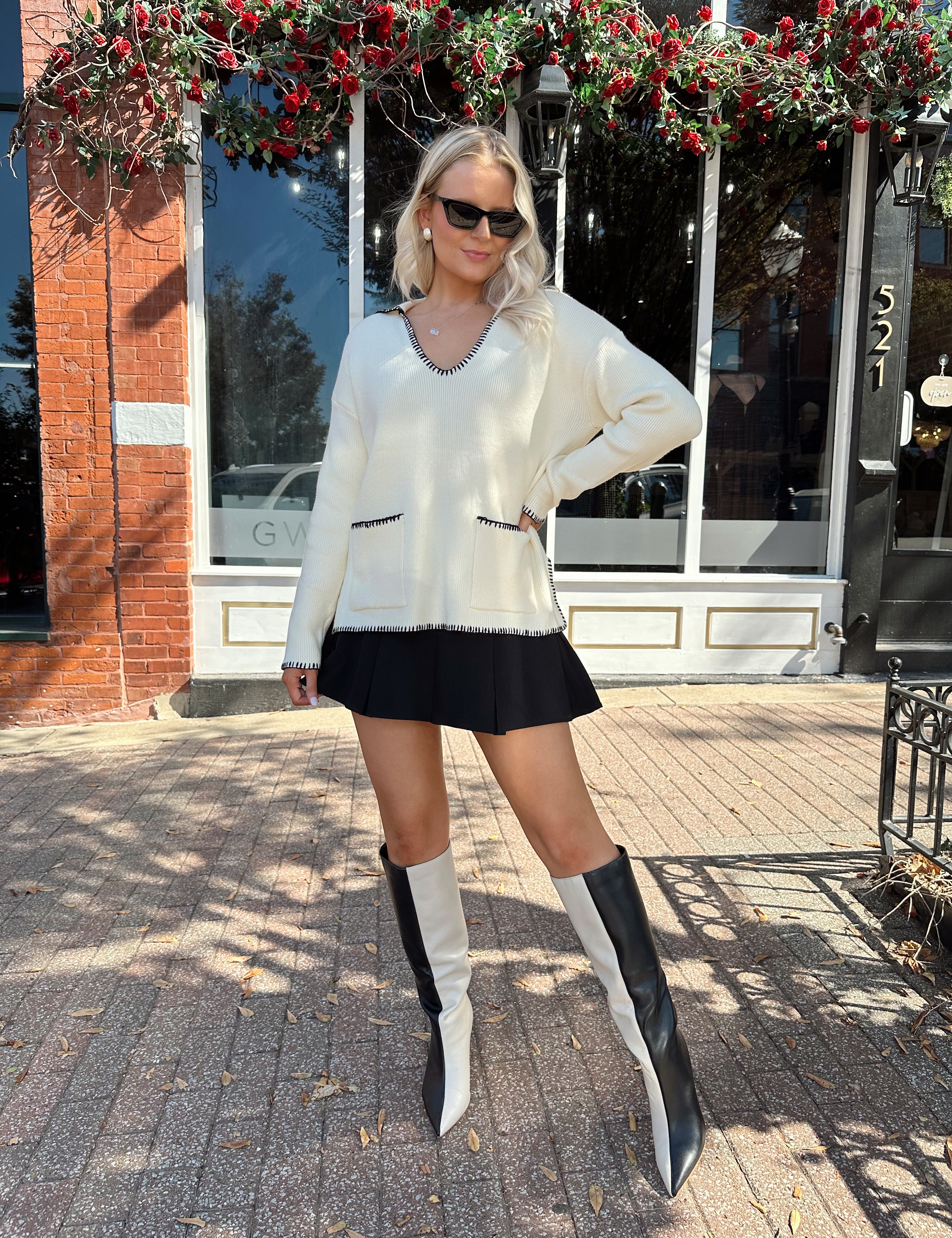 Woman wearing ivory sweater with black stitch detailing and front pockets. 