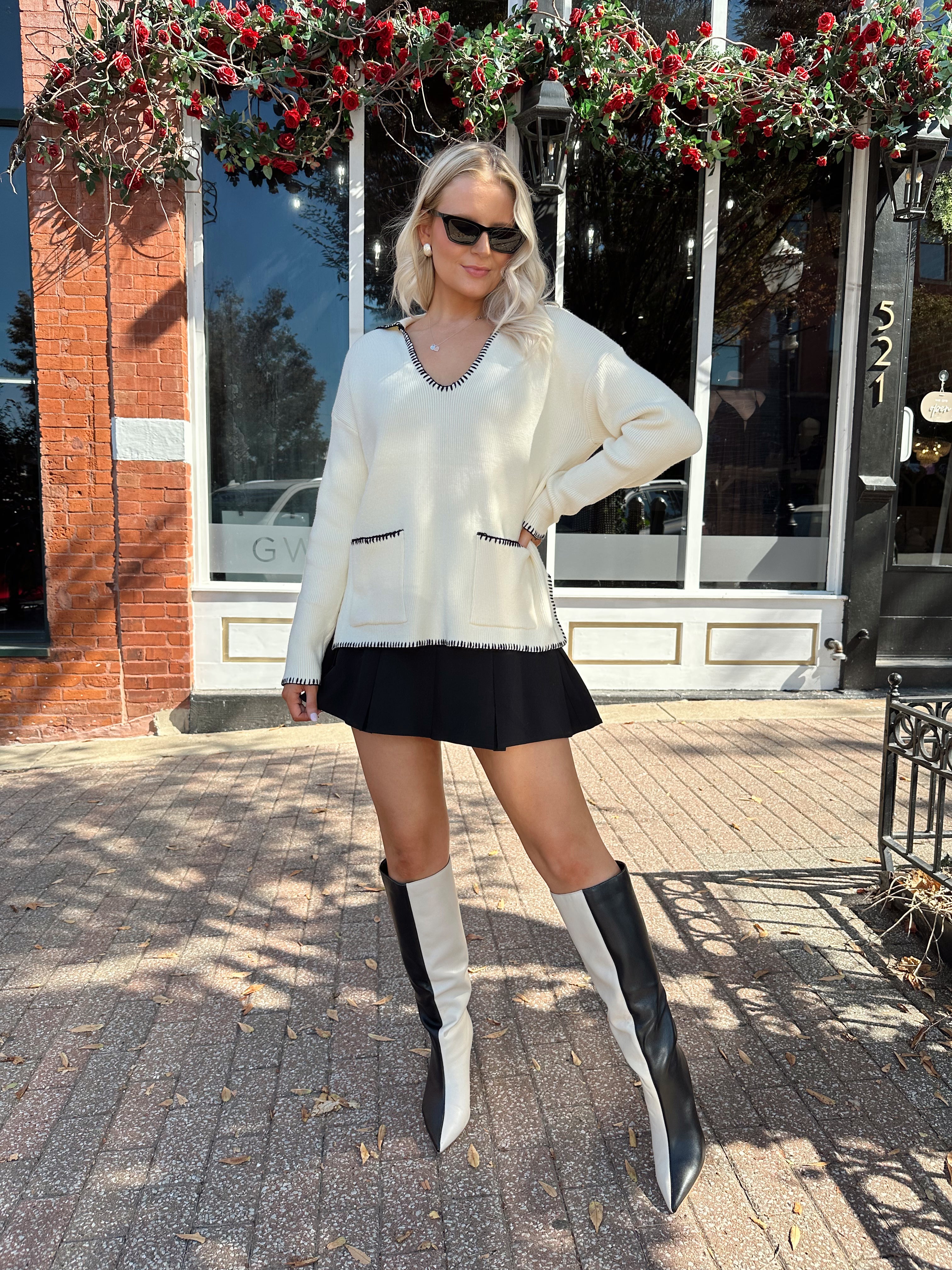 Woman wearing ivory sweater with black stitch detailing and front pockets. 