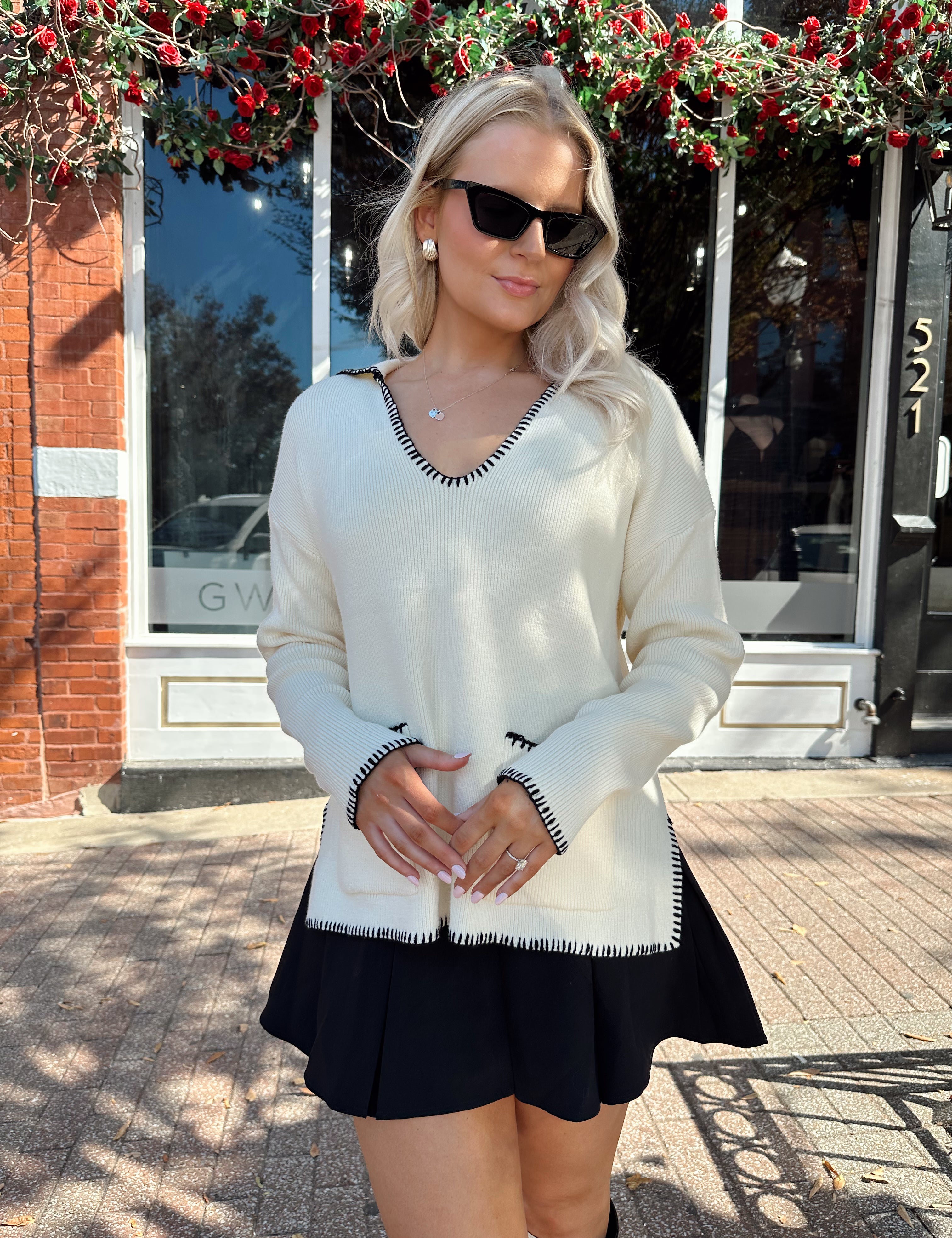Woman wearing ivory sweater with black stitch detailing and front pockets. 