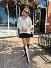 Woman wearing ivory sweater with black stitch detailing and front pockets. 