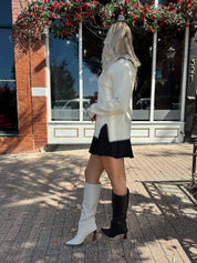 Woman wearing ivory sweater with black stitch detailing and front pockets. 