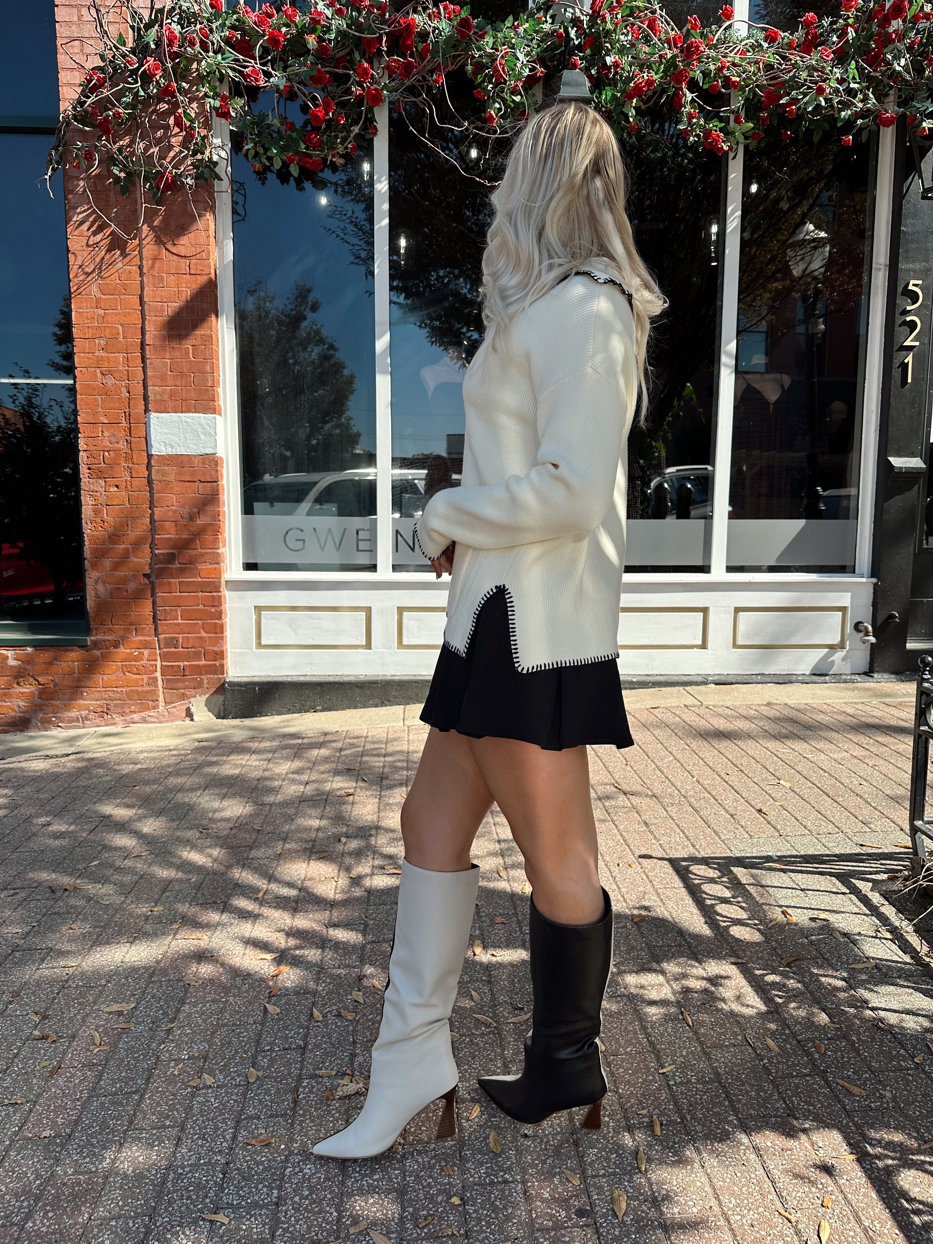 Woman wearing ivory sweater with black stitch detailing and front pockets. 
