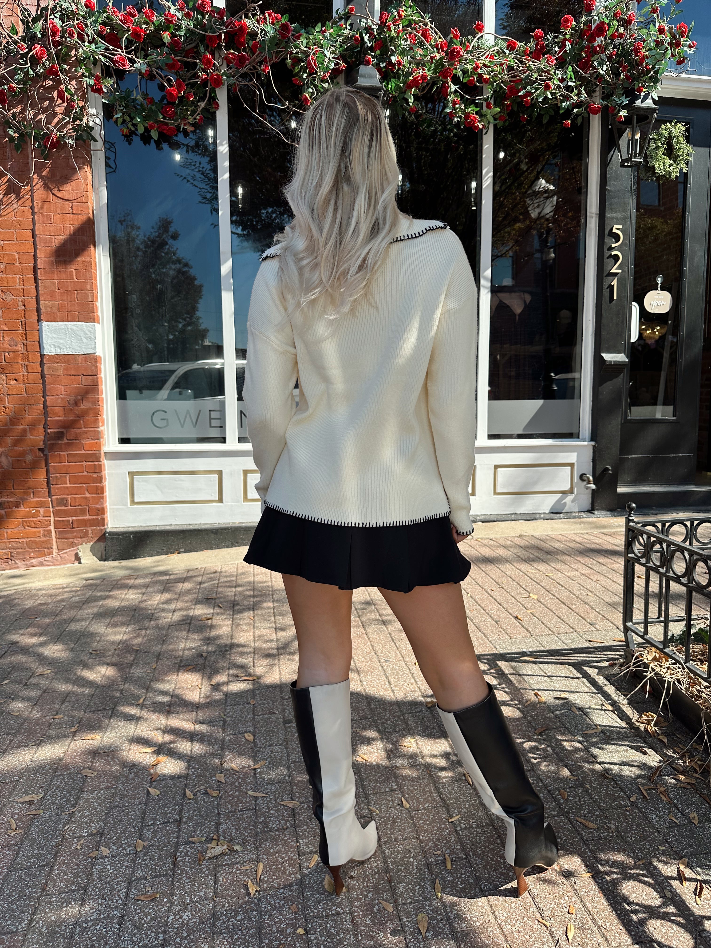 Woman wearing ivory sweater with black stitch detailing and front pockets. 