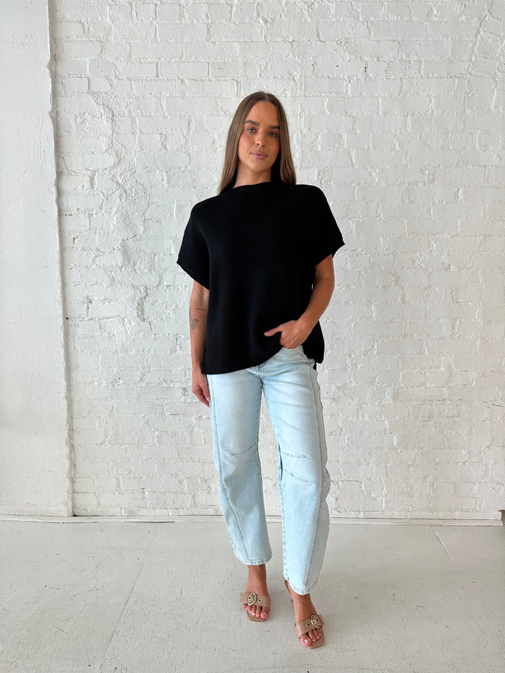 Woman wearing an oversized black knit short sleeve sweater top with light wash barrel jean. 