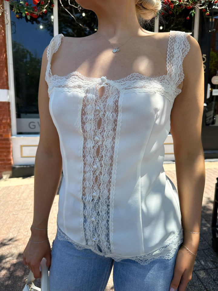 Woman wearing cream tank top with lace trim, and button detailing with a tie back.  