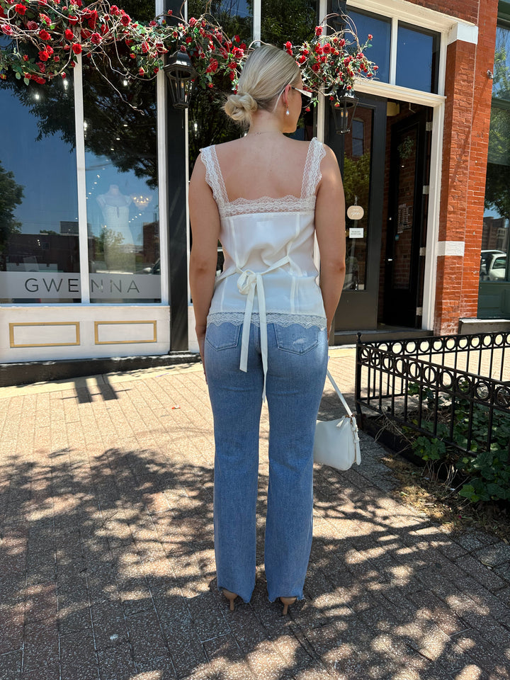Woman wearing cream tank top with lace trim, and button detailing with a tie back. 