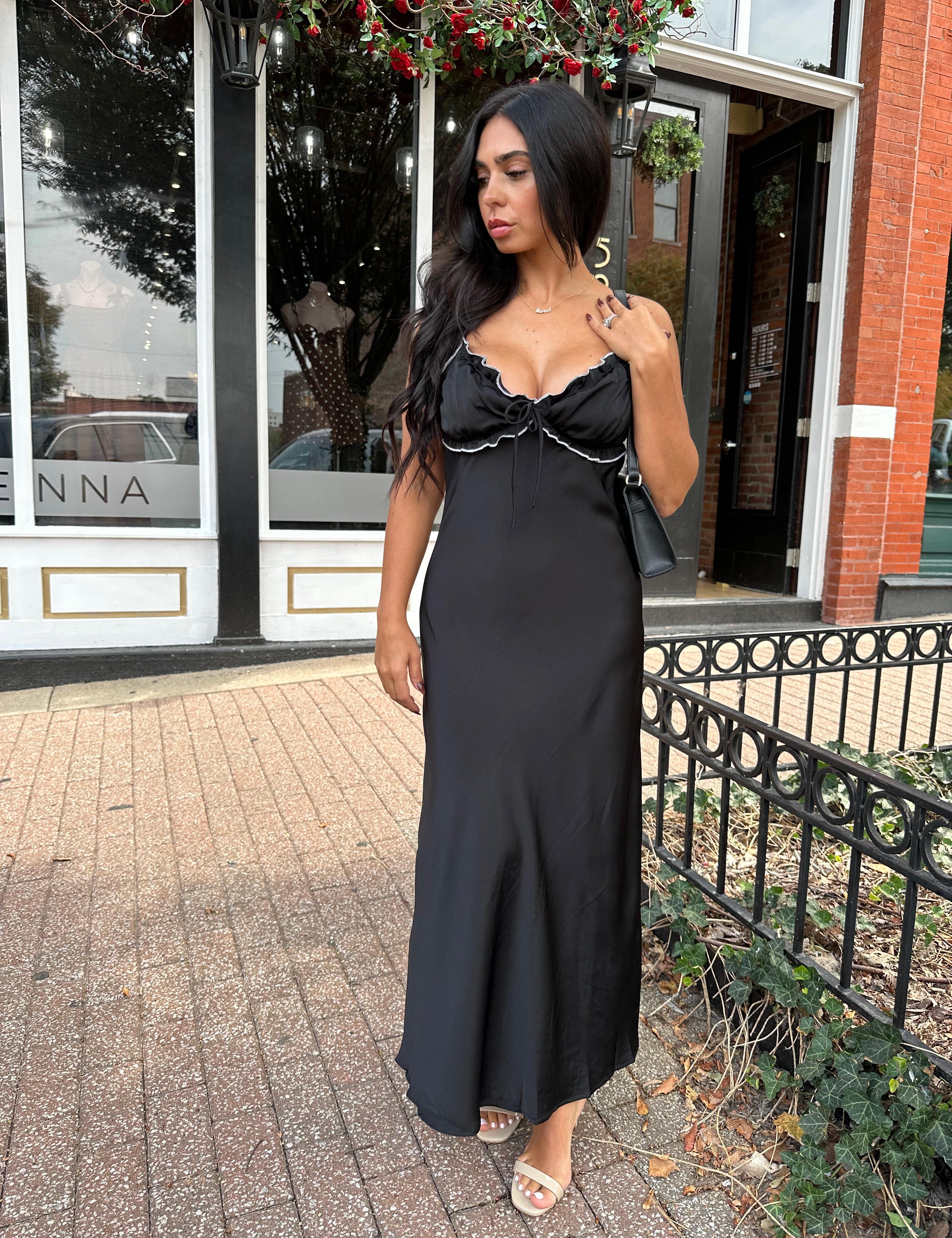 Woman wearing black satin maxi dress with white ruffle trim and open back. 
