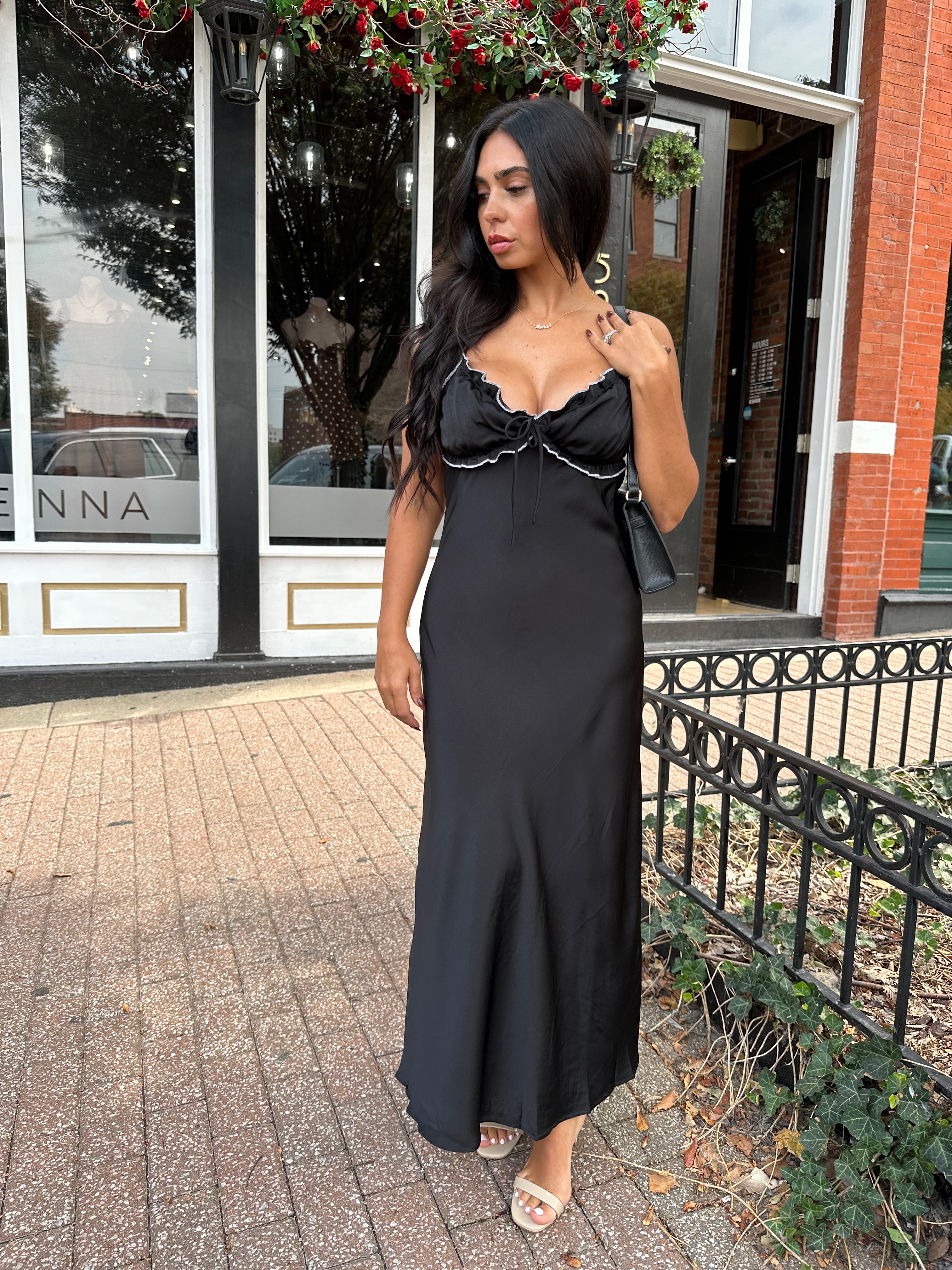 Woman wearing black satin maxi dress with white ruffle trim and open back. 