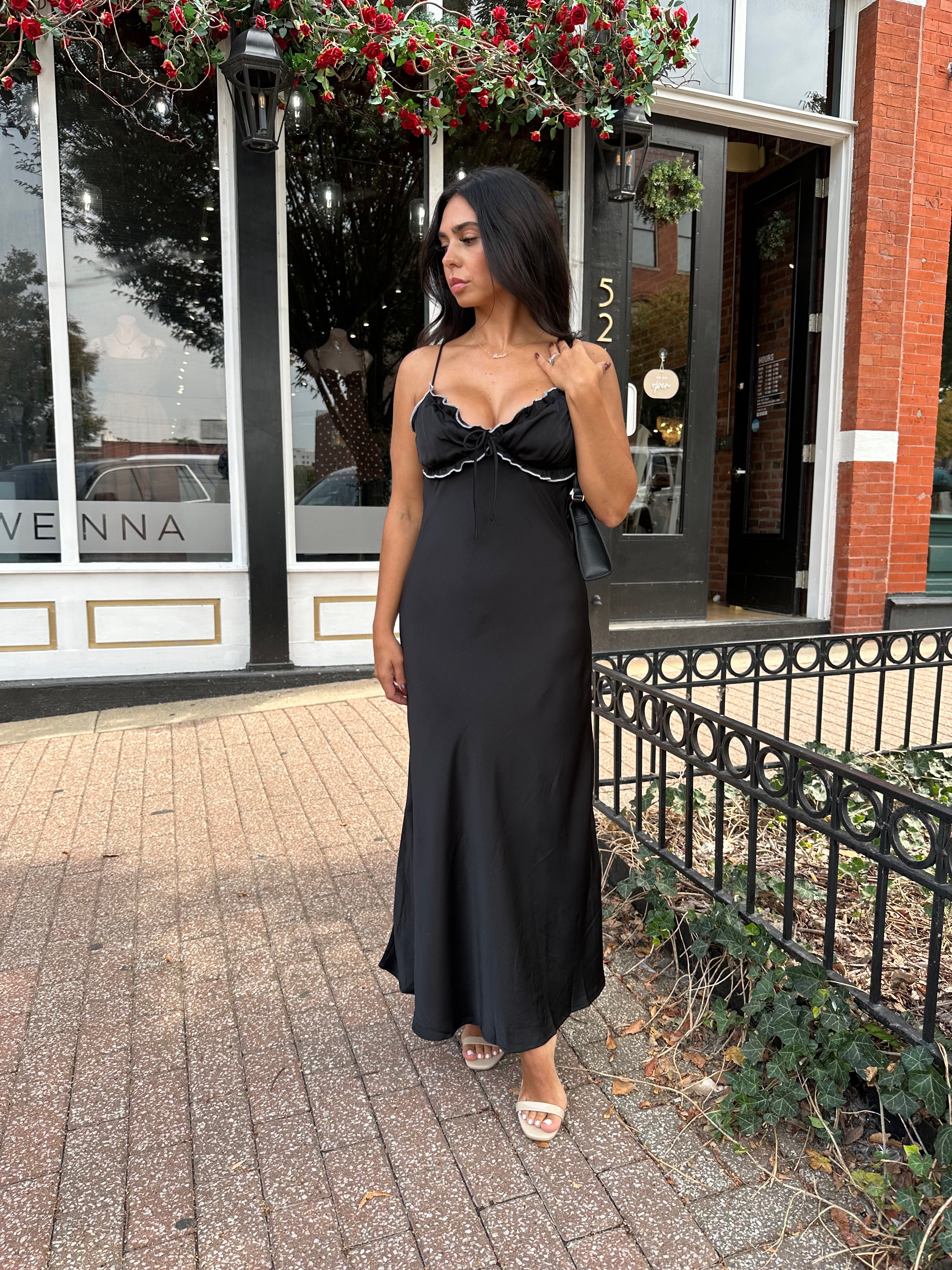 Woman wearing black satin maxi dress with white ruffle trim and open back. 