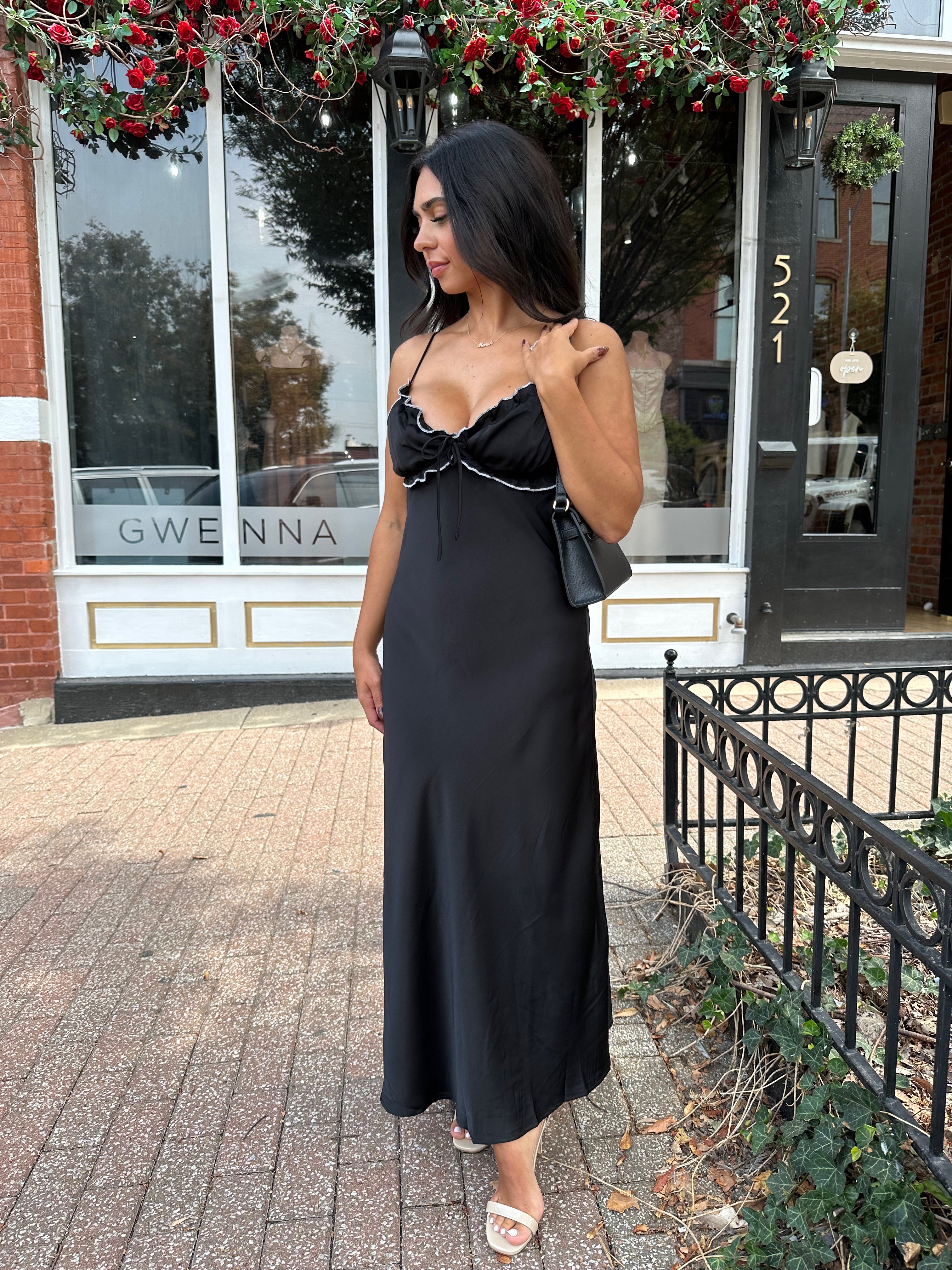 Woman wearing black satin maxi dress with white ruffle trim and open back. 
