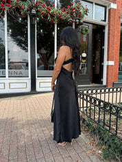  Woman wearing black satin maxi dress with white ruffle trim and open back. 