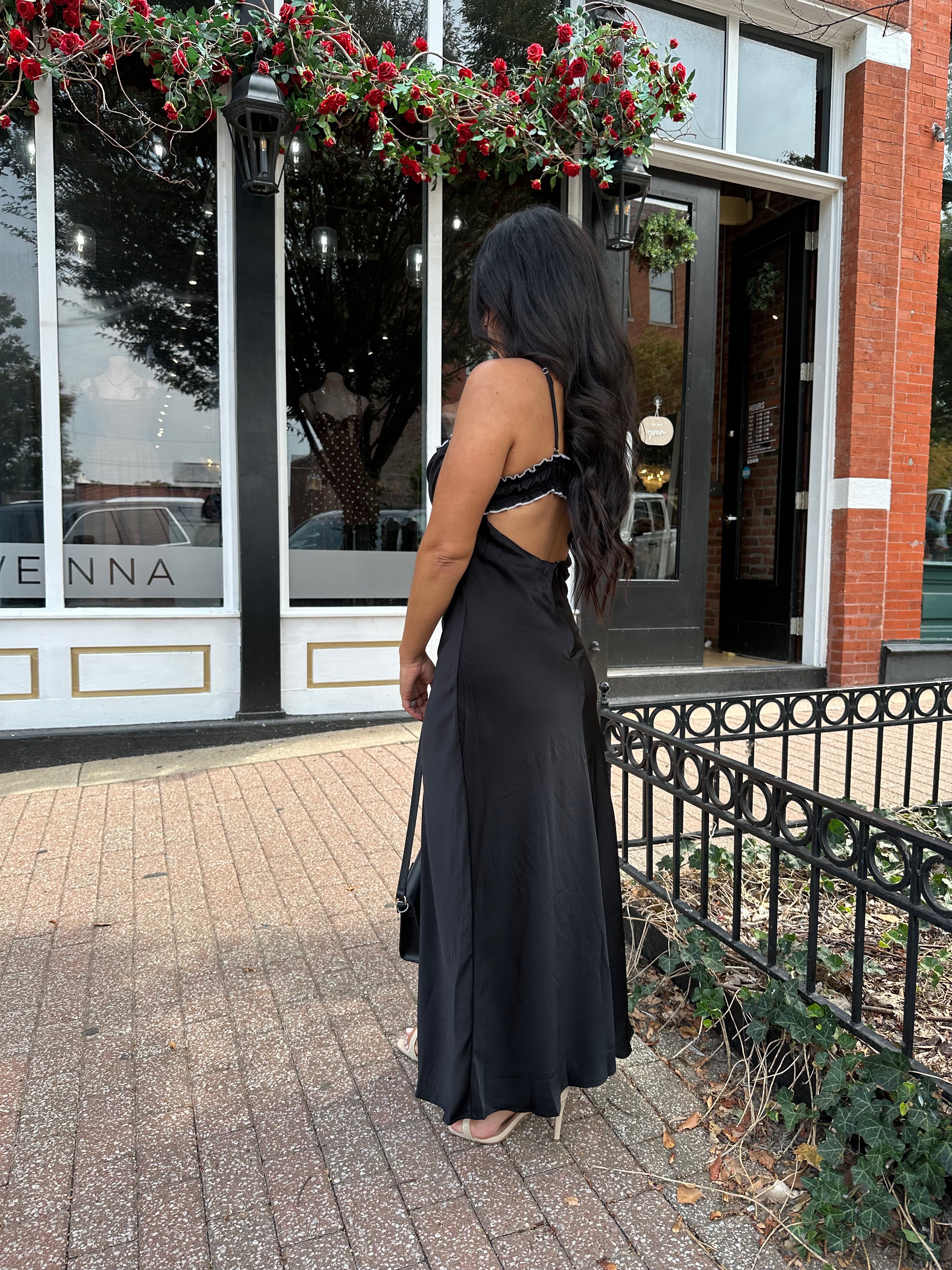  Woman wearing black satin maxi dress with white ruffle trim and open back. 