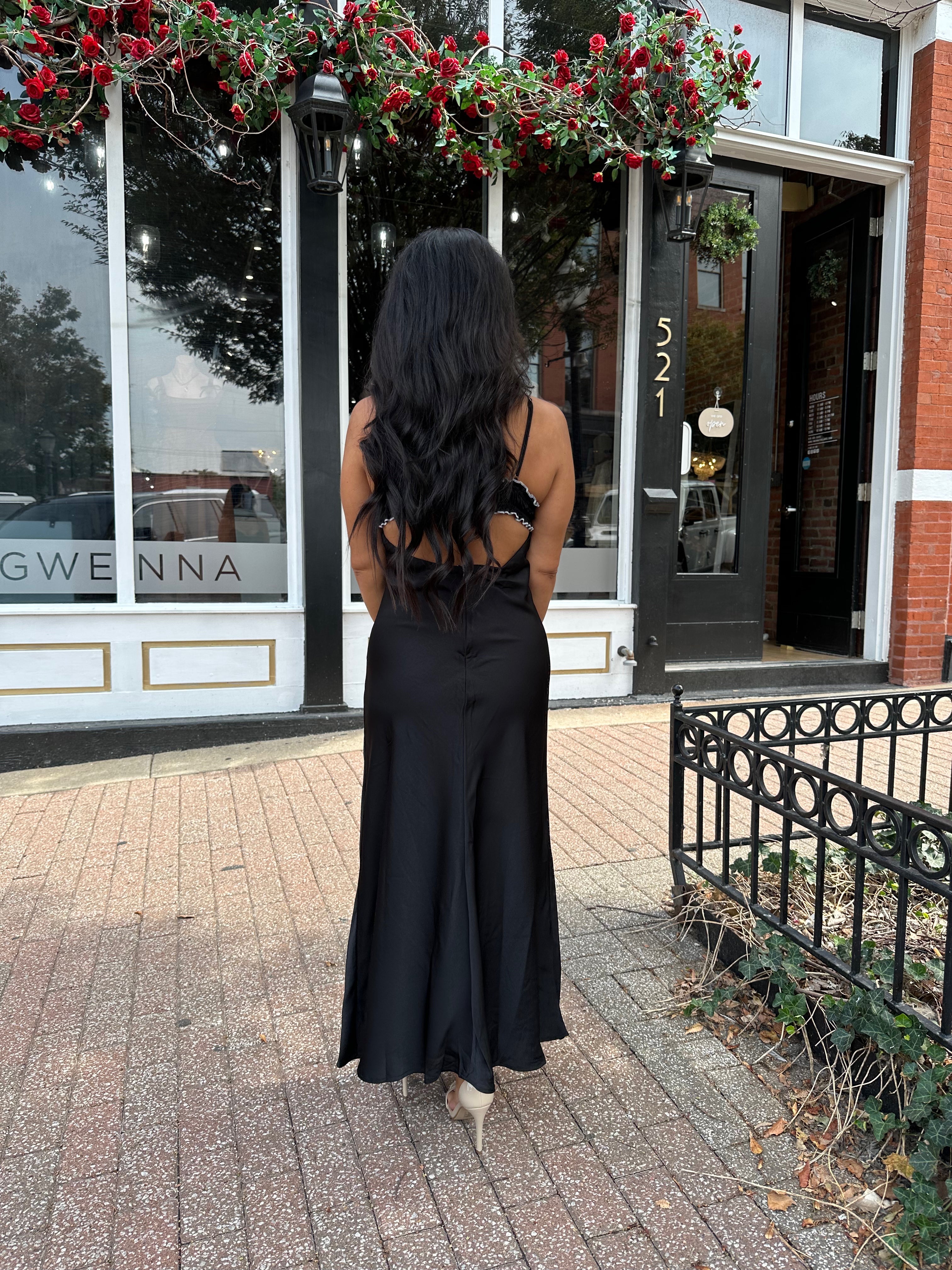 Woman wearing black satin maxi dress with white ruffle trim and open back. 