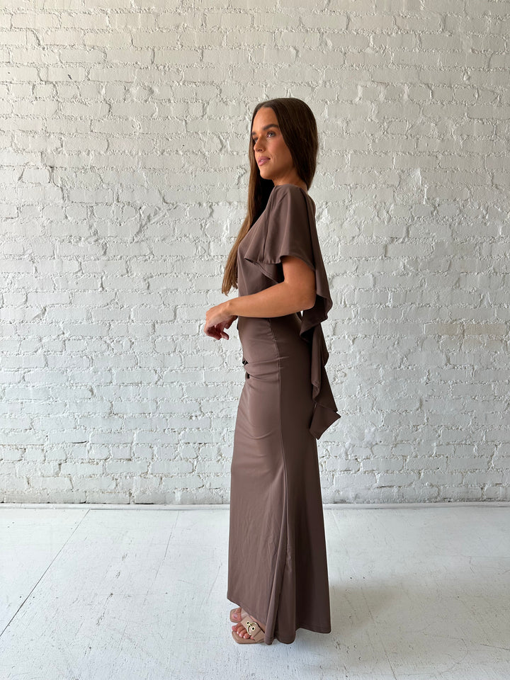 Woman wearing brown maxi dress with open back and flowy draped sleeves. 