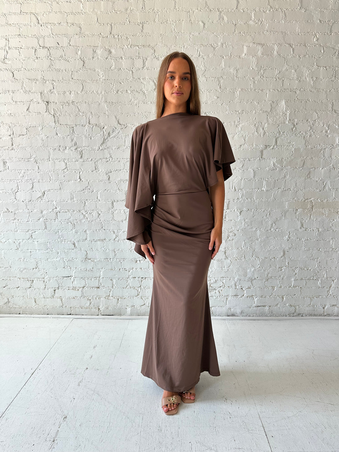 Woman wearing brown maxi dress with open back and flowy draped sleeves. 