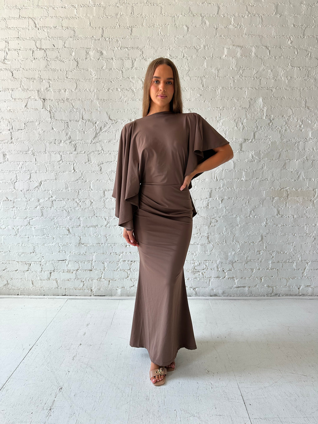 Woman wearing brown maxi dress with open back and flowy draped sleeves. 