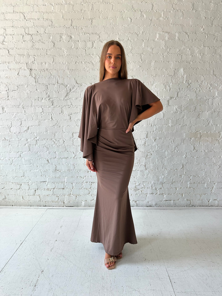 Woman wearing brown maxi dress with open back and flowy draped sleeves. 